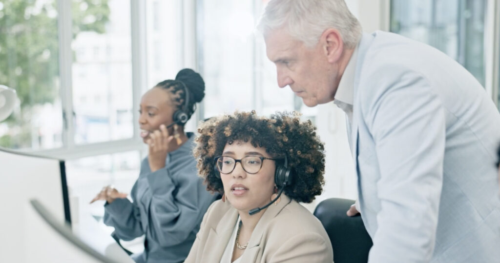 Sales manager coaching a headset-wearing employee at her desk while another team member works nearby in an office.